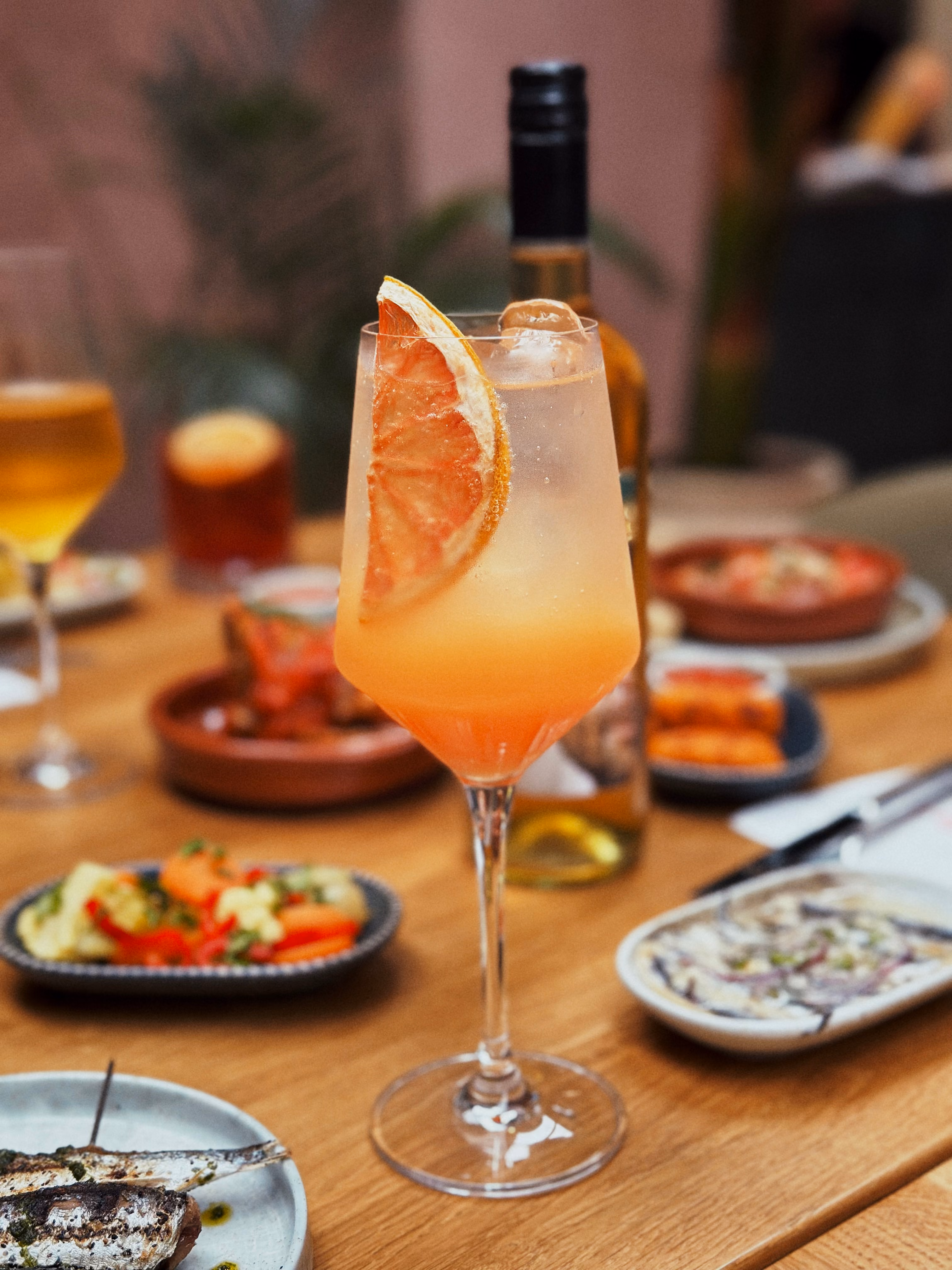 Orange cocktail with grapefruit slice garnish on a wooden table surrounded by small plates of food.