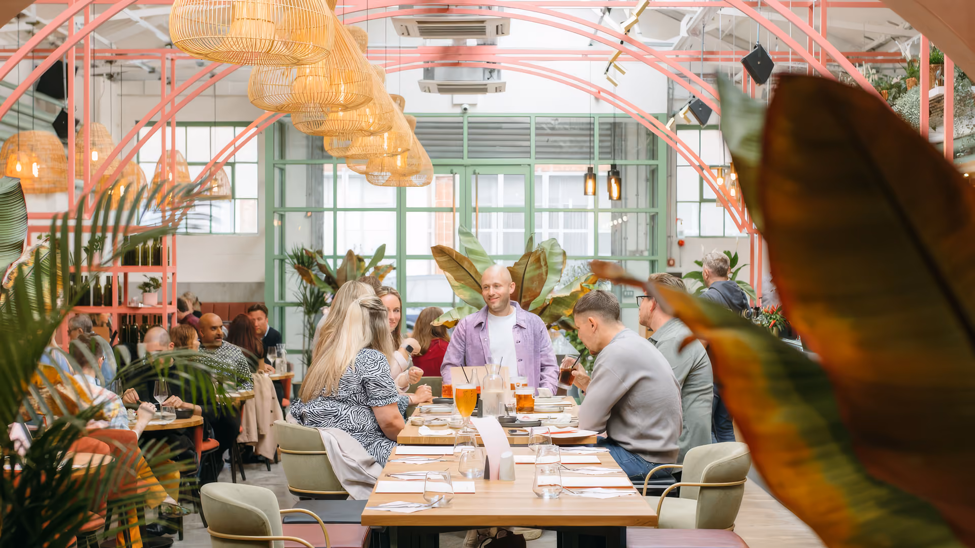 People dining and socializing at a table in a bright, plant-filled restaurant with pink arches.