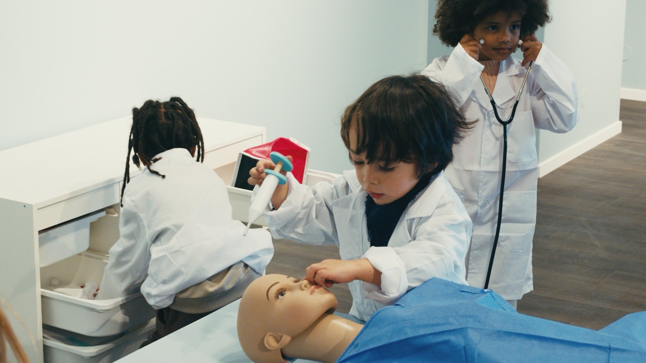 Le Village de Noé - Trois enfants déguisés en médecins jouent avec un mannequin d'entraînement, l'un tenant une seringue jouet et un autre utilisant un stéthoscope.