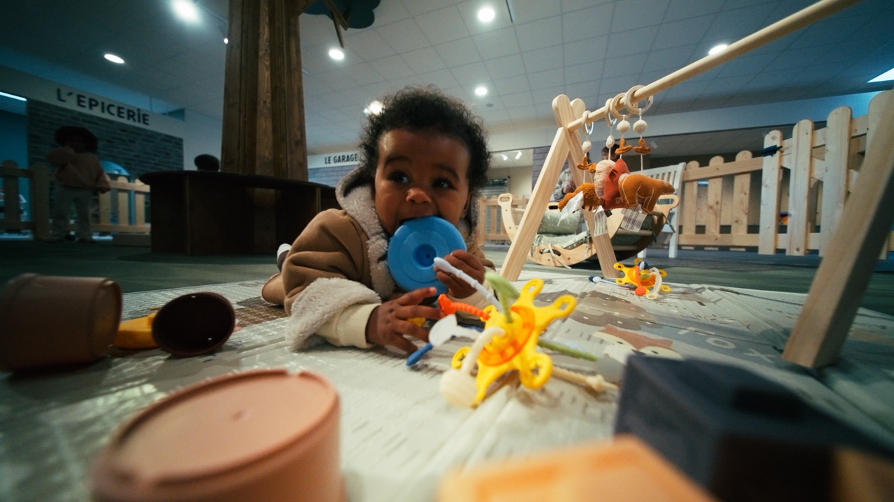 Le Village de Noé - Bébé allongé sur un tapis de jeu mordant un jouet bleu, entouré de jouets colorés dans un espace intérieur avec barrières en bois.