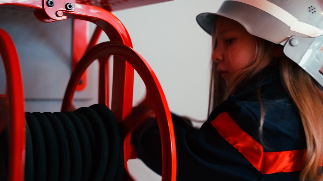 Le Village de Noé - Jeune fille portant un casque de pompier et un uniforme en train de manipuler un tuyau d'incendie enroulé sur un support rouge.