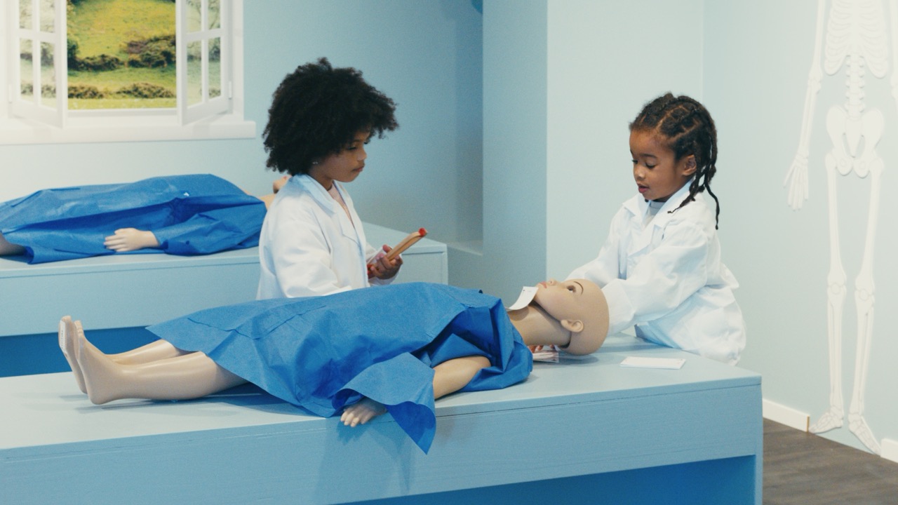 Le Village de Noé - Deux enfants en blouses blanches jouent à examiner des mannequins médicaux dans une salle de classe aux murs bleus.