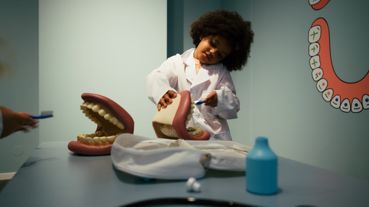 Le Village de Noé - Une enfant en blouse blanche brosse un modèle géant de dents dans une salle avec une illustration de dents sur le mur.