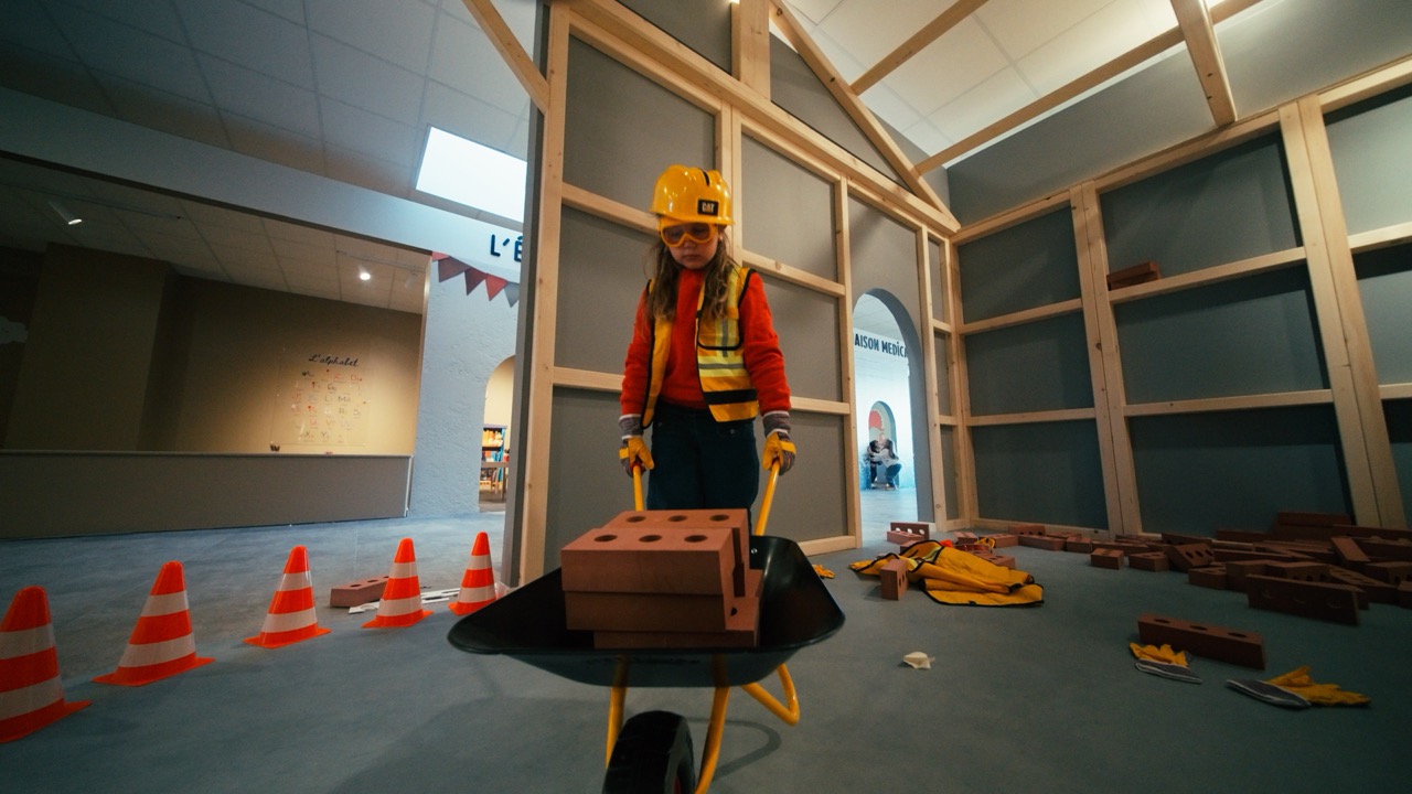 Le Village de Noé - Enfant portant un casque, des lunettes de protection, un gilet réfléchissant et des gants, poussant une brouette avec des briques dans une salle de jeu d'imitation de chantier.