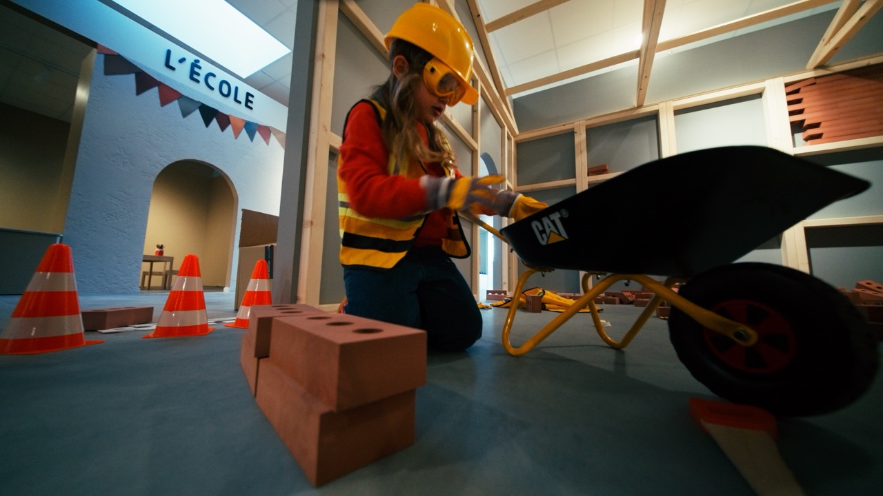 Le Village de Noé - Enfant en tenue de chantier avec casque et gilets de sécurité jouant avec une brouette et des briques dans une salle d'activités intitulée 'L'École'.