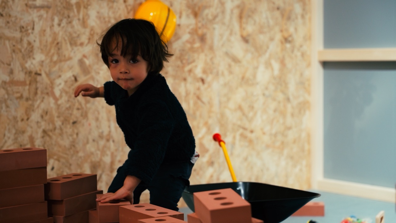 Le Village de Noé - Un enfant joue avec des briques en plastique à l'intérieur, avec une brouette noire et un casque jaune en arrière-plan.