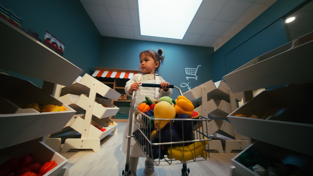 Le Village de Noé - Une petite fille poussant un chariot de courses rempli de légumes et fruits jouets dans un magasin simulé pour enfants.