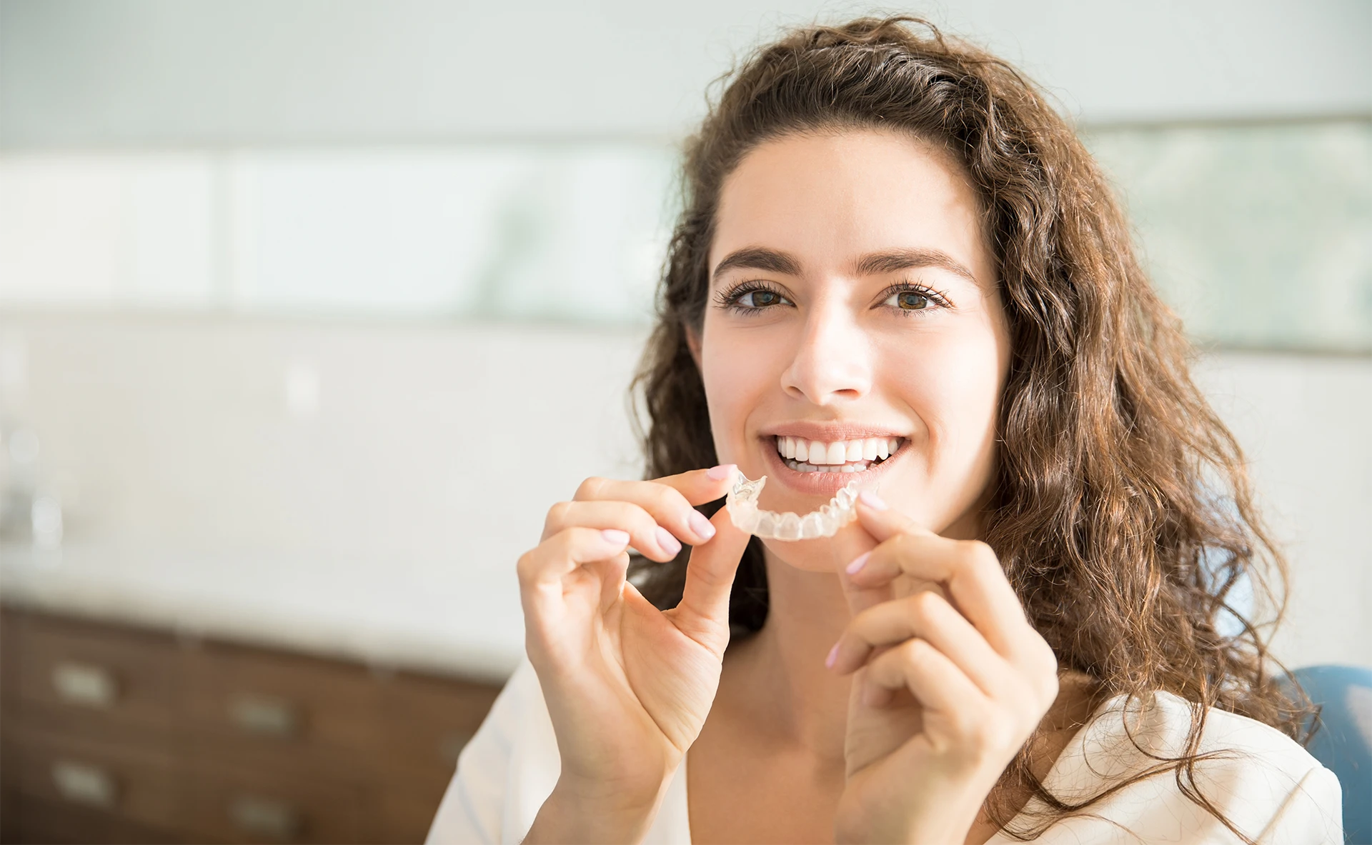 Smiling woman holding a clear dental aligner in a bright indoor setting.