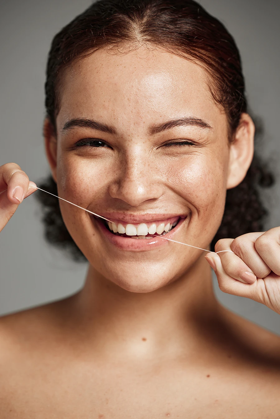 Smiling woman flossing her teeth with dental floss against a neutral background.