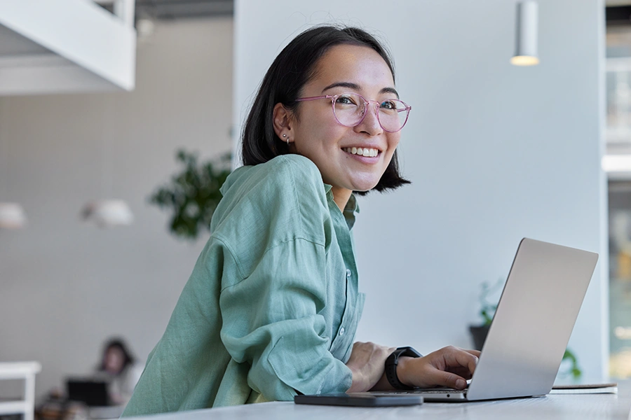 Smiling woman wearing glasses and a green shirt working on a laptop at a bright workspace.
