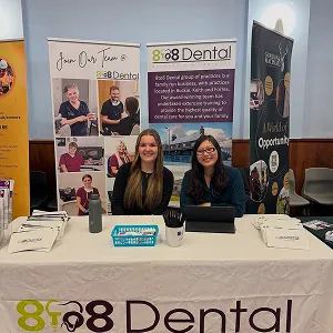 Two women sitting at a 8to8 Dental information booth with banners and promotional materials on the table indoors.