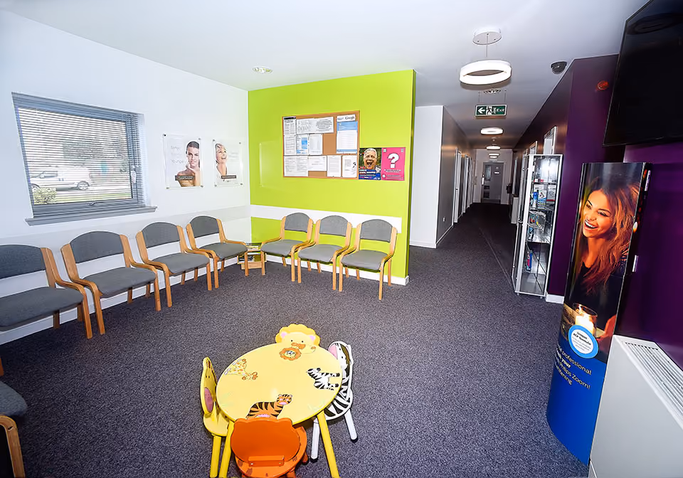 Bright waiting room with gray chairs along the walls, a colorful children's table with animal designs, a green accent wall with a bulletin board, and a hallway leading to multiple doors.