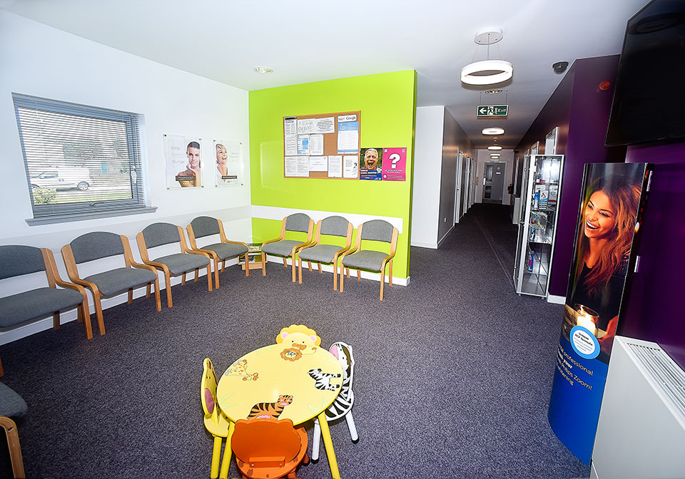 Bright waiting room with gray chairs along the walls, a colorful children's table with animal designs, a green accent wall with a bulletin board, and a hallway leading to multiple doors.