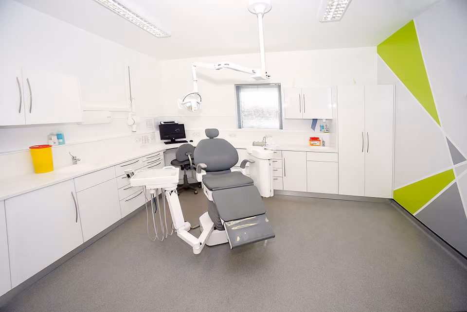 Modern dental clinic room with a gray dental chair, overhead light, white cabinets, and a gray floor.
