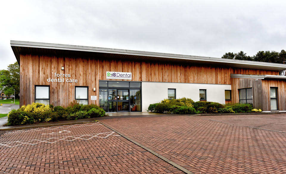 Modern building with wooden paneling and a sign reading 'forres dental care' and '888 Dental' above the entrance with a brick parking lot in front.