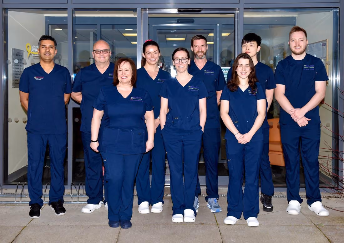 Group of nine healthcare professionals wearing navy blue scrubs standing in front of glass doors, smiling at the camera.