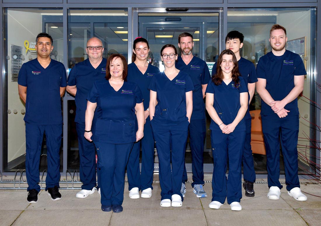 Group of nine healthcare professionals wearing navy blue scrubs standing in front of glass doors, smiling at the camera.