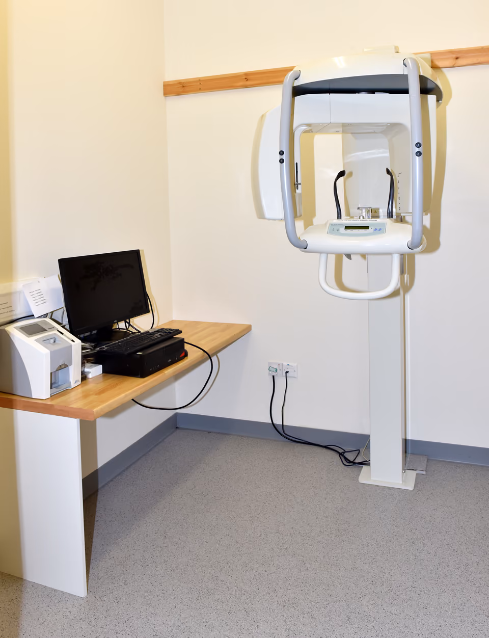 Dental panoramic X-ray machine next to a desk with a computer and printer in a clinical room.