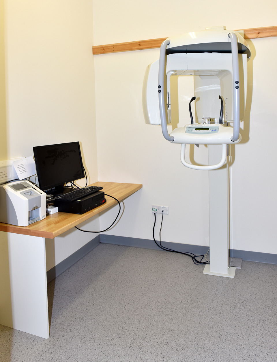 Dental panoramic X-ray machine next to a desk with a computer and printer in a clinical room.