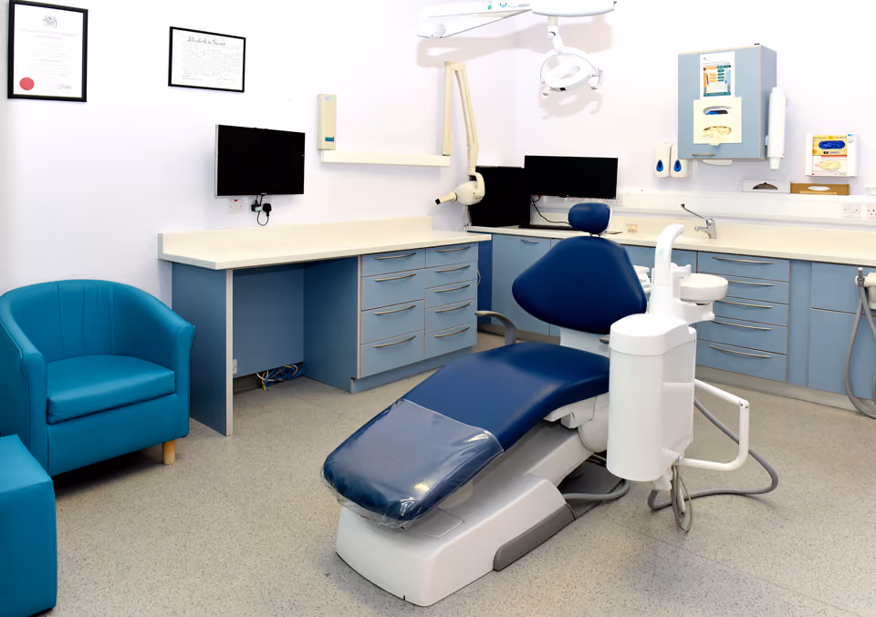 Modern dental office with blue dental chair, overhead light, computer monitors, blue cabinets, and teal armchair.