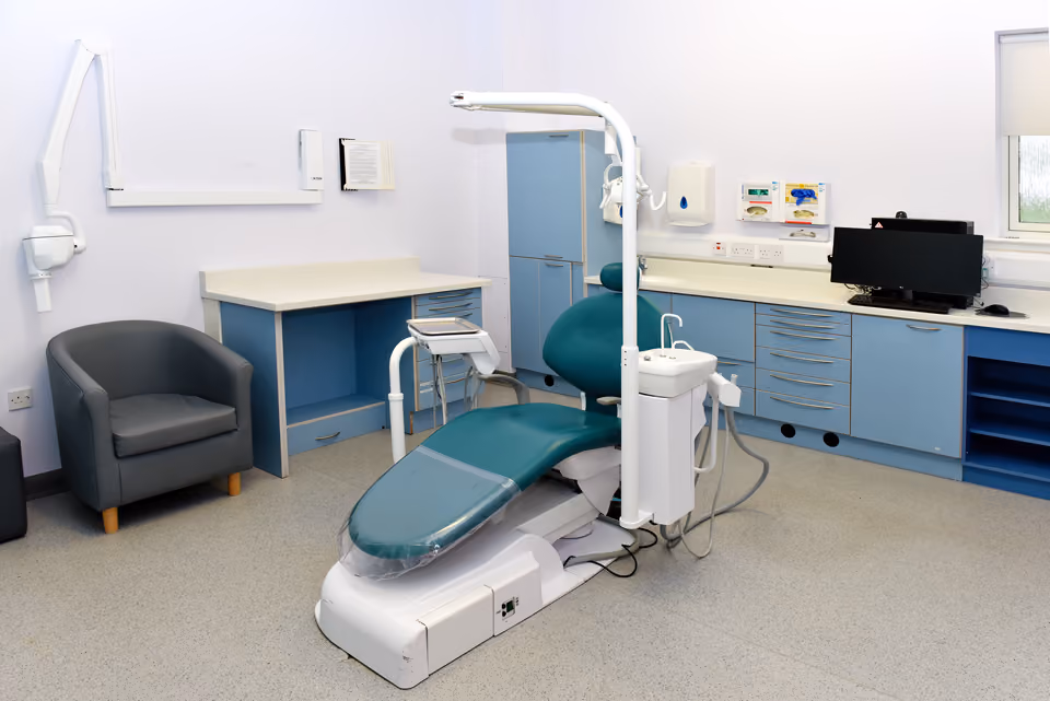 Empty dental examination room with a teal dental chair, blue cabinets, countertop, computer, and a gray armchair.