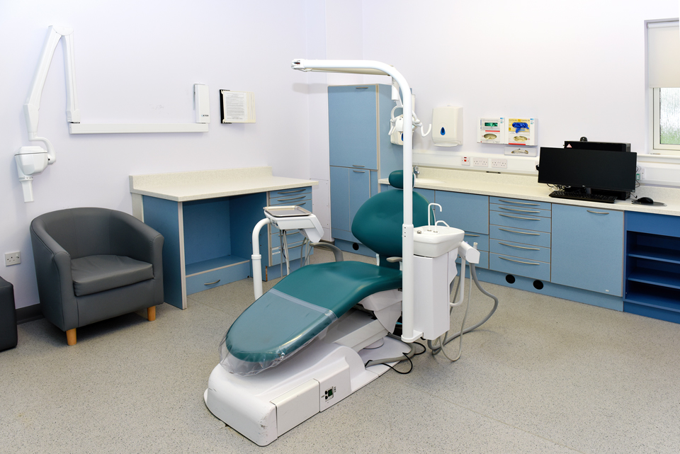 Empty dental examination room with a teal dental chair, blue cabinets, countertop, computer, and a gray armchair.