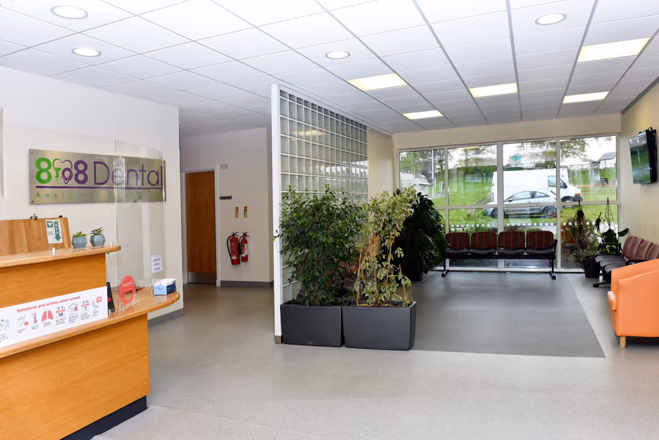 Bright dental clinic waiting area featuring a wooden reception desk with protective screen, potted plants, rows of seating, and large windows overlooking parked cars outside.