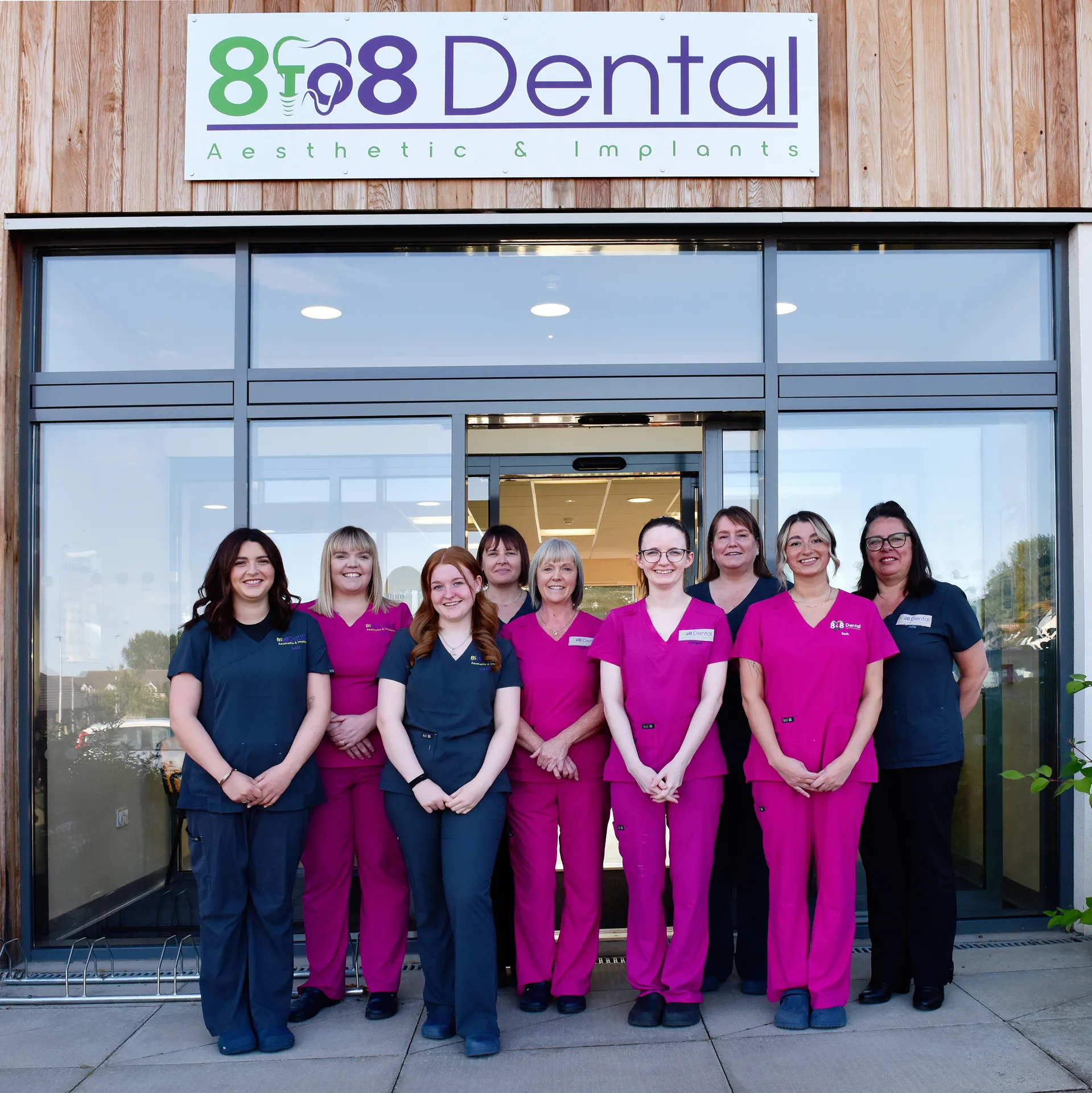 Group of nine dental staff in blue and pink scrubs standing outside 808 Dental clinic under the clinic sign.