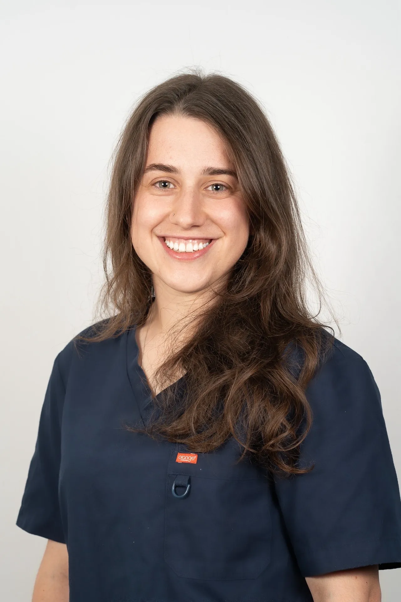 Smiling young woman with long brown hair wearing a navy blue medical scrub top against a plain white background.