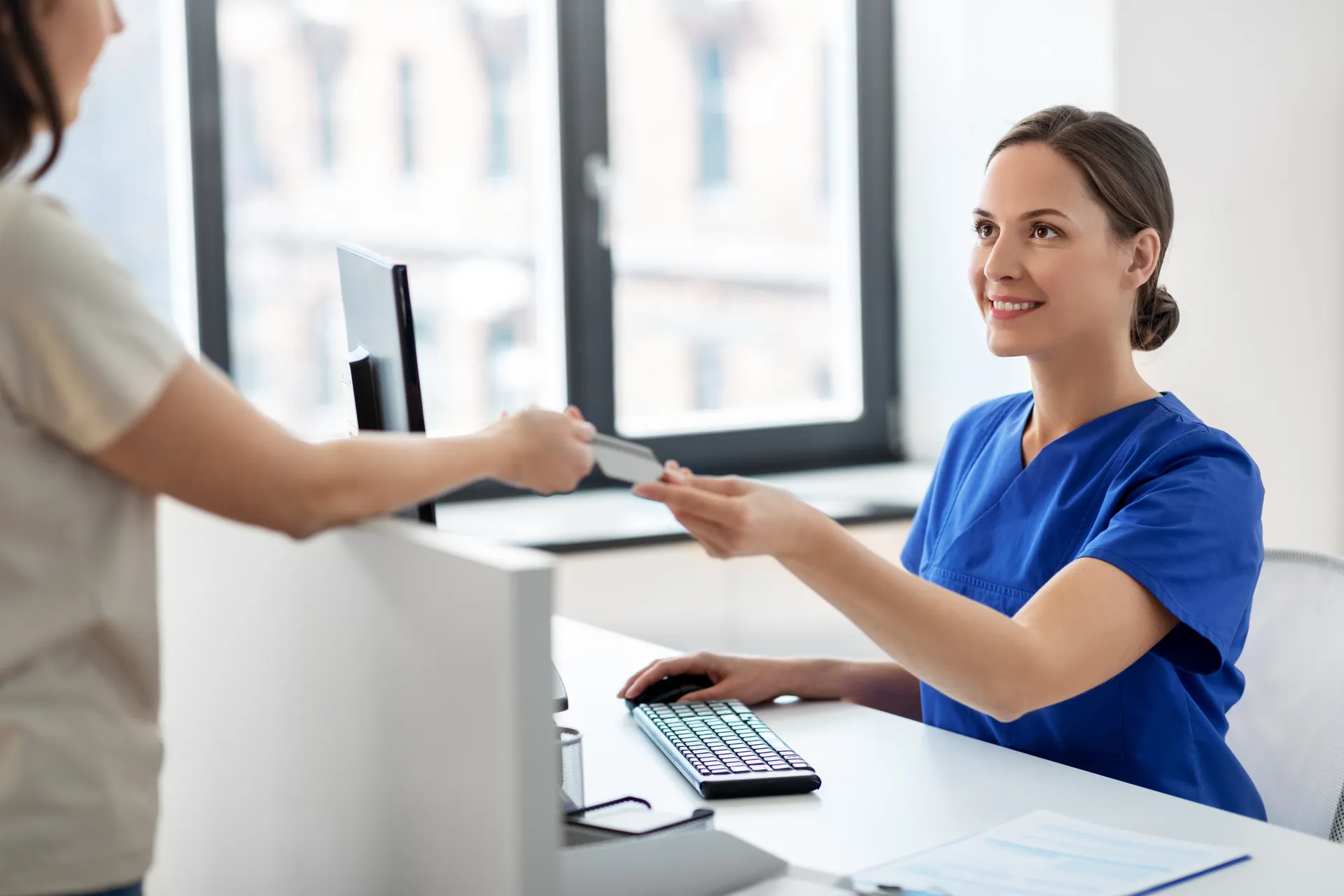 Smiling female nurse in blue scrubs receiving a card from a patient at a reception desk.