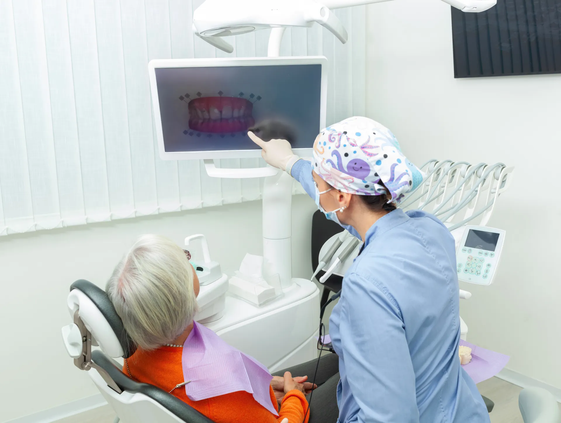 Dentist in blue scrubs and mask showing dental X-ray on screen to elderly patient seated in dental chair.