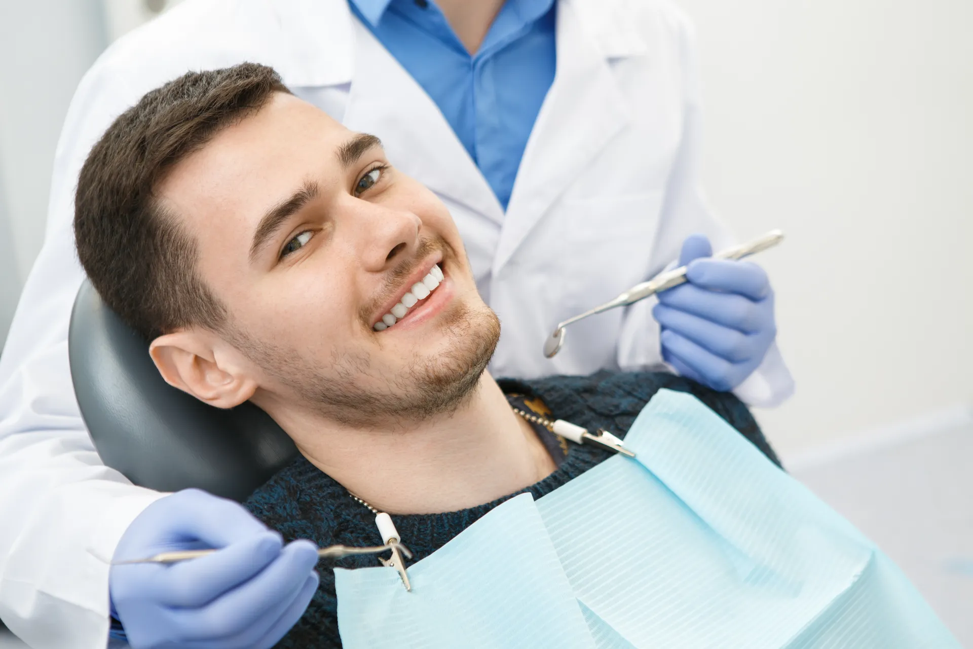 Smiling young man reclined in a dental chair with a dentist holding dental instruments nearby.