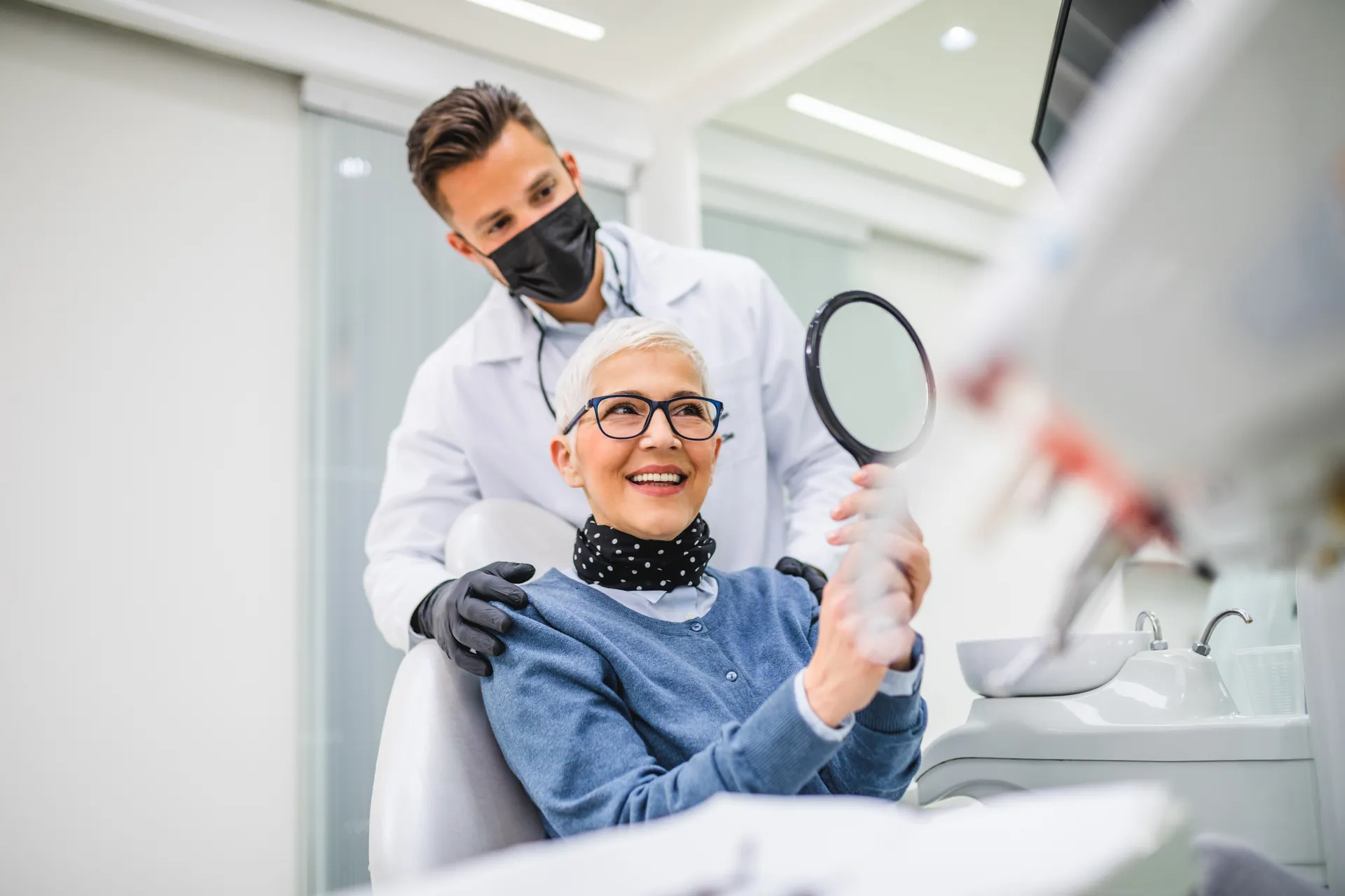 Smiling mature woman holding a hand mirror while a male dentist wearing a mask stands behind her in a dental clinic.