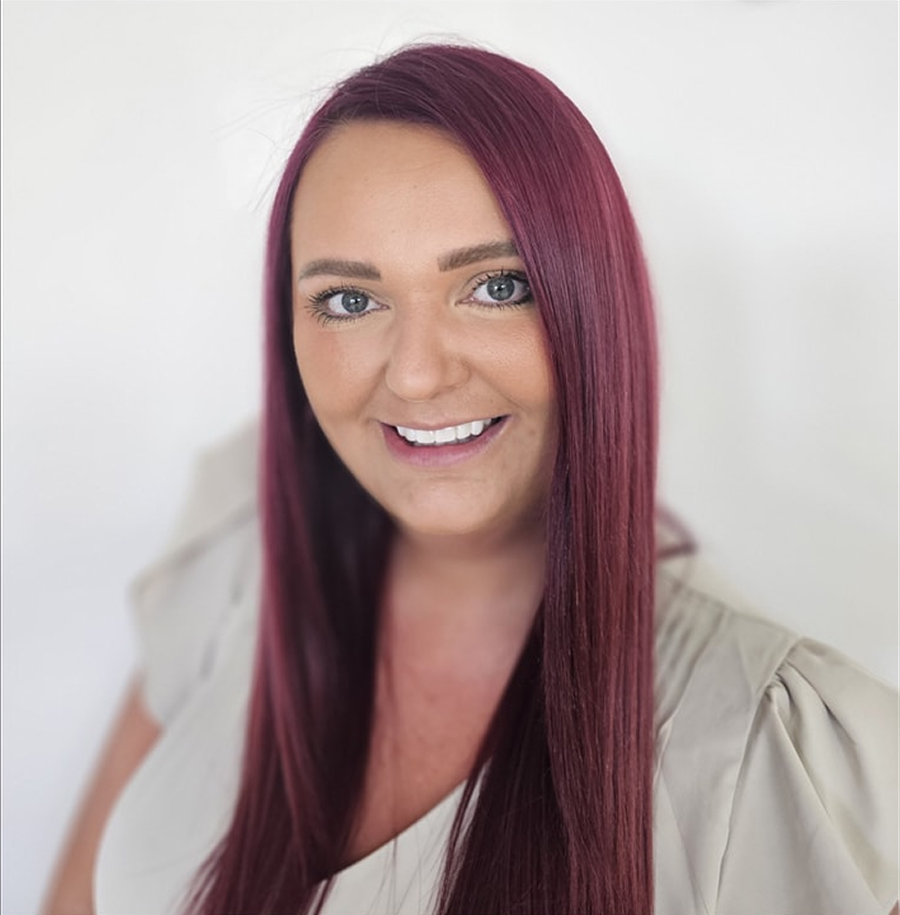Smiling woman with long straight burgundy hair wearing a light-colored top against a plain white background.
