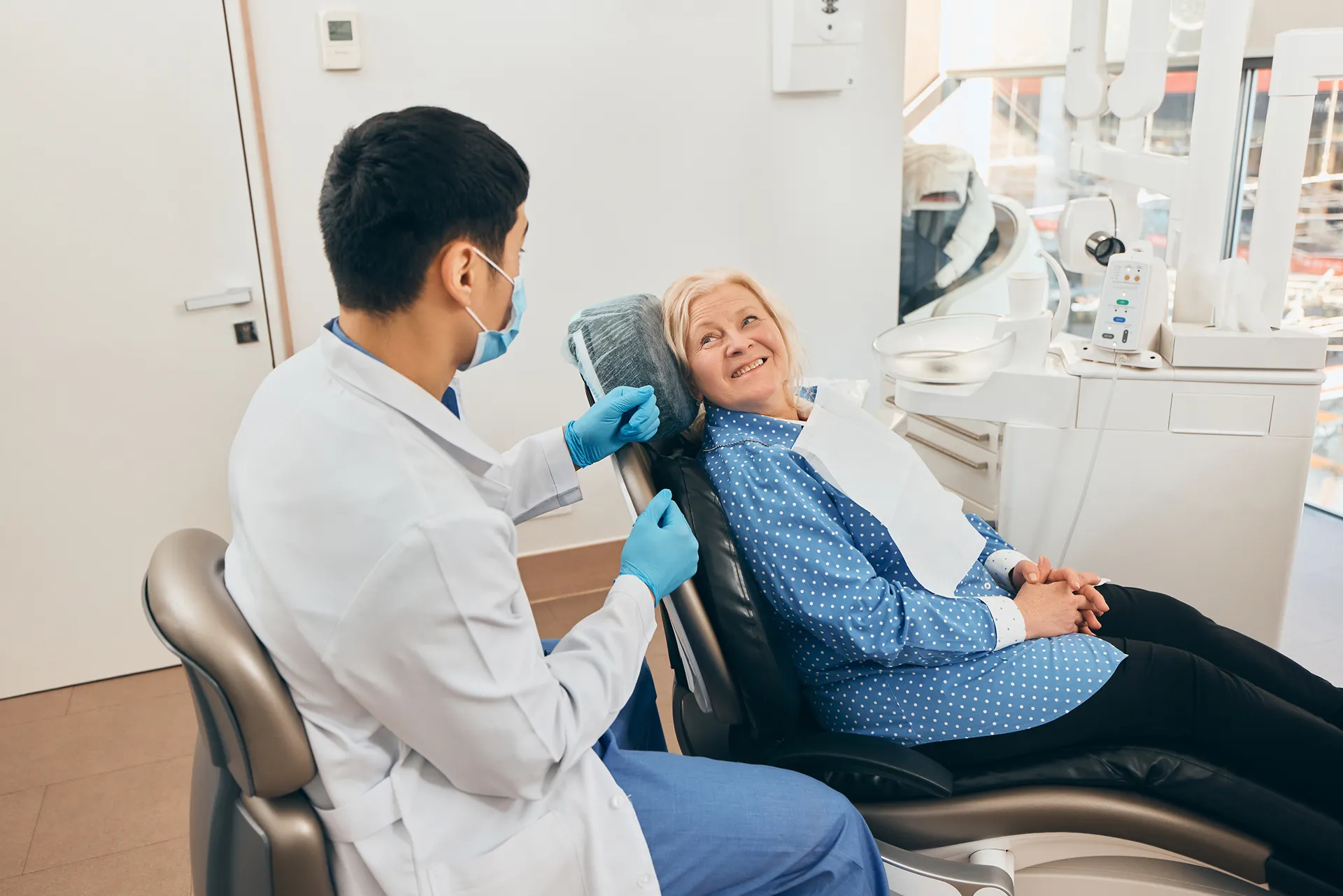 Dentist in a white coat and mask talking to a smiling elderly woman seated in a dental chair.
