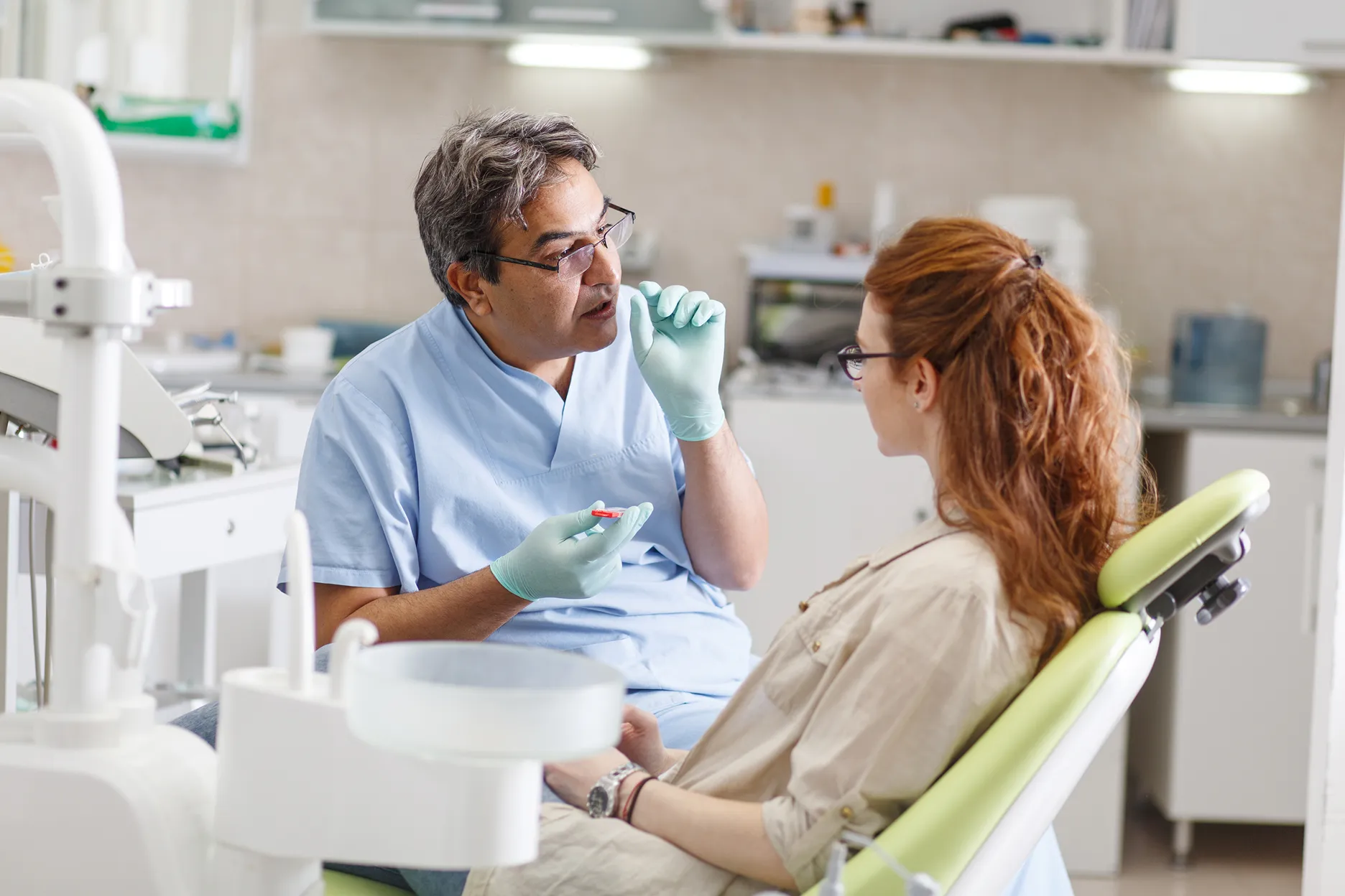 Dentist wearing blue scrubs and gloves explaining a dental product to a woman seated in a green dental chair.