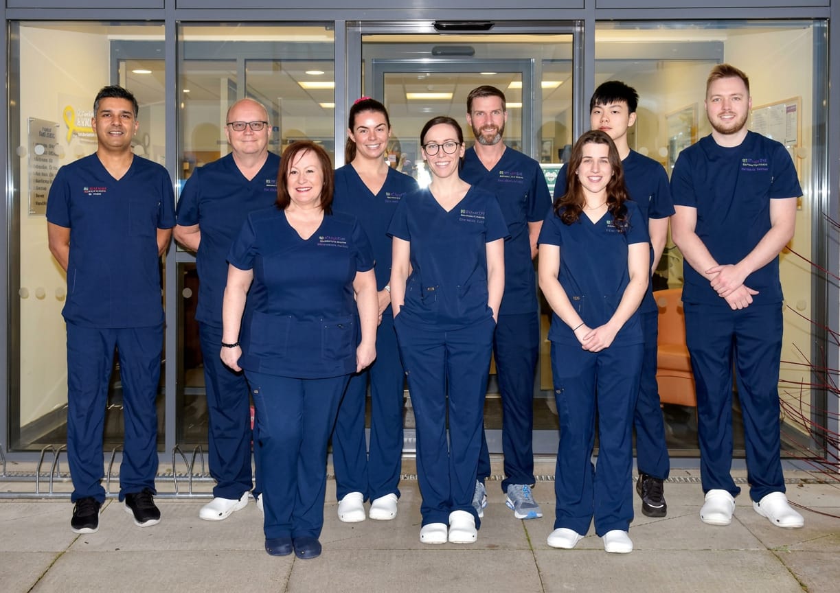 Group of nine healthcare professionals in navy blue scrubs standing and smiling in front of glass doors of a medical facility.
