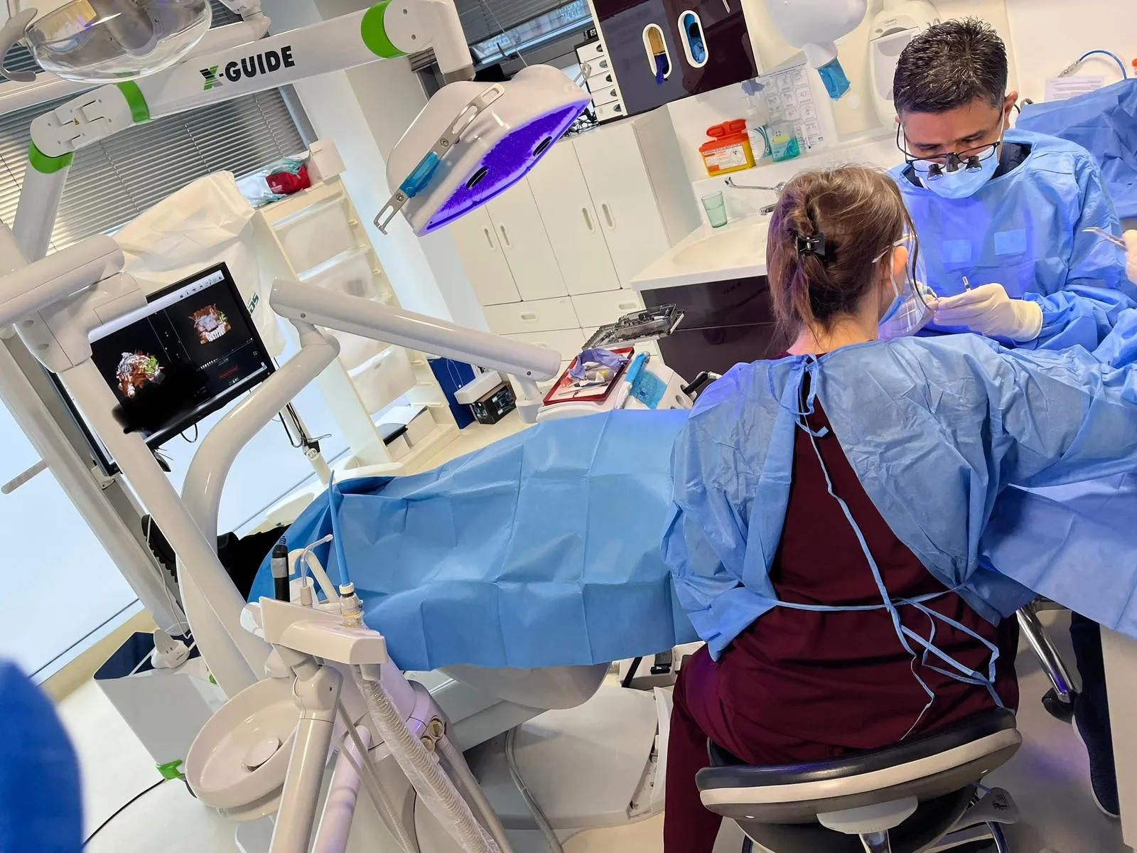 Dentist in magnifying eyewear performing dental procedure on patient with assistant in a modern dental clinic.