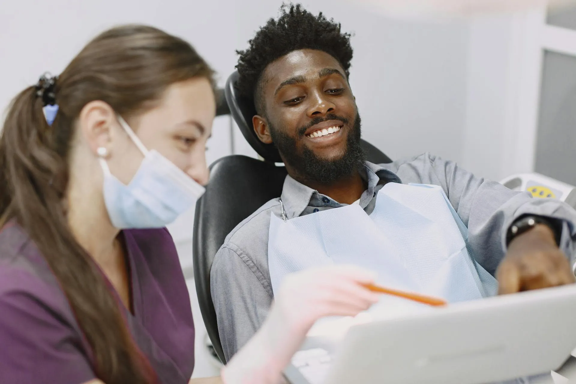 Smiling man in dental chair reviews information with masked dentist holding a pen in clinic.
