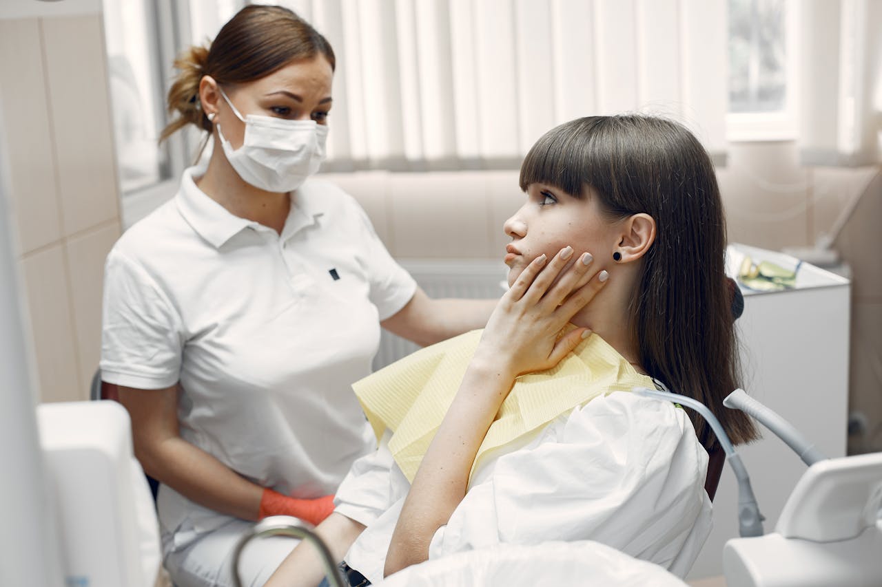 Young woman patient touching her jaw while dentist in mask speaks to her in dental clinic.