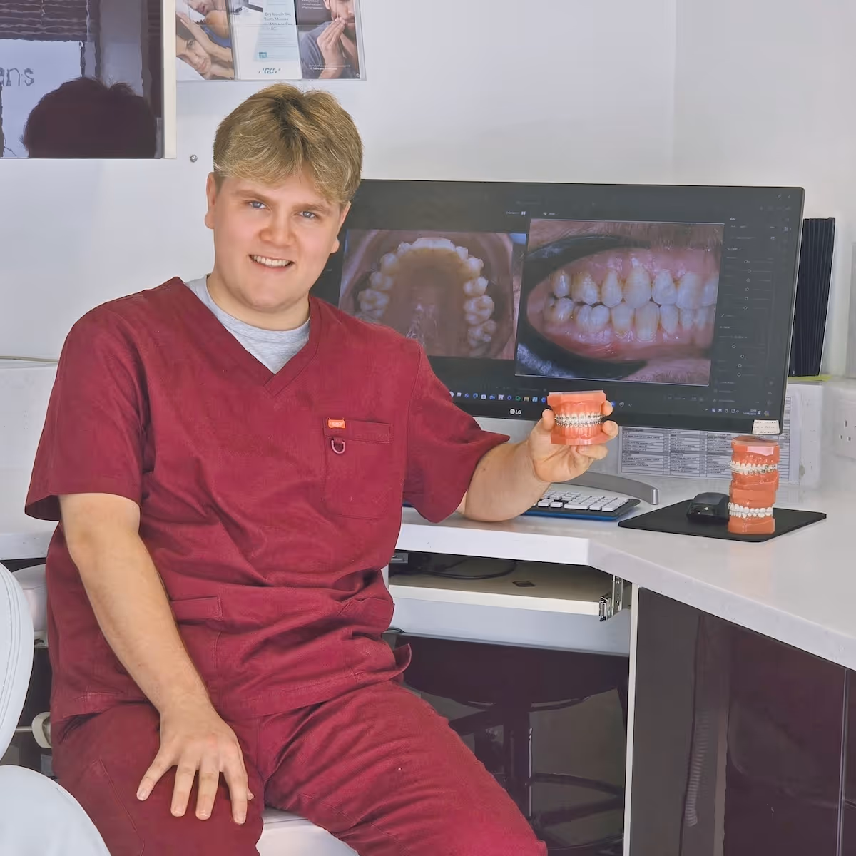 Dentist in red scrubs holding dental model with braces in a clinic with dental images on screen.