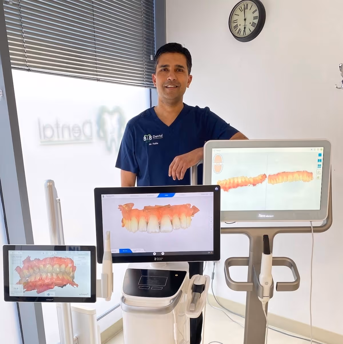Male dentist in blue scrubs standing behind dental scanners showing 3D tooth images on screens.