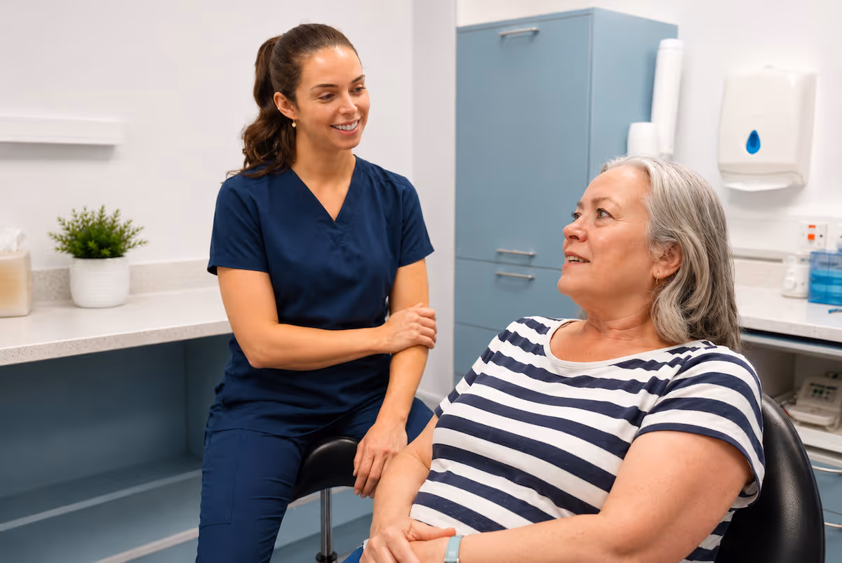Smiling female healthcare worker in blue scrubs talking to an elderly woman in a clinic room.