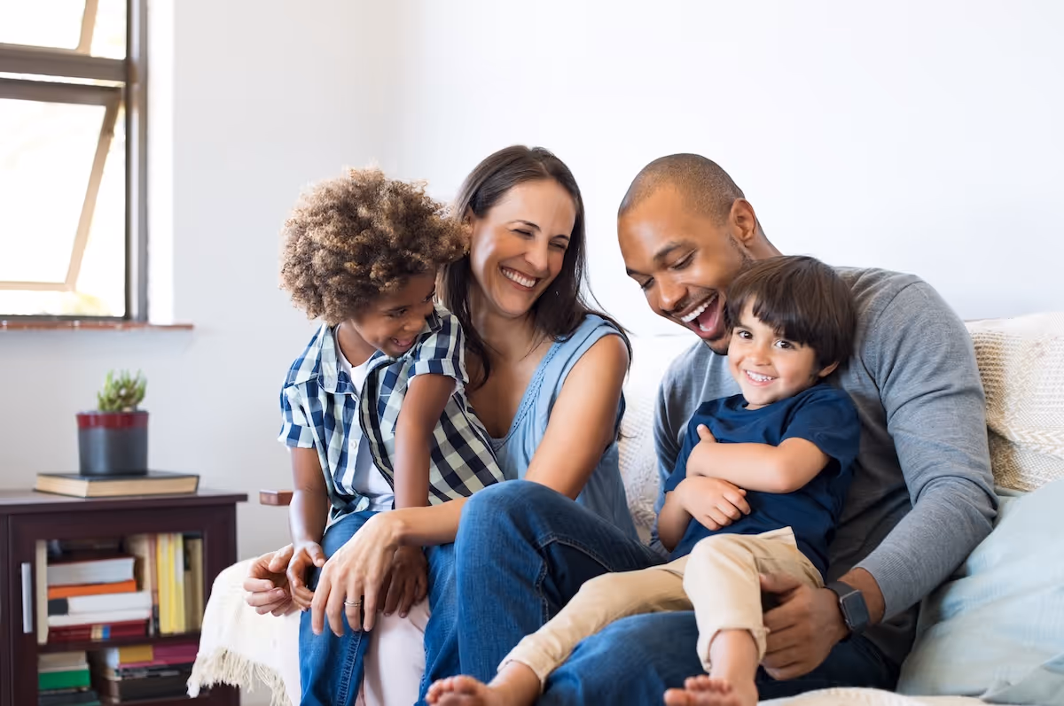 Smiling family of four sitting closely on a couch in a bright room with a window and bookshelf.