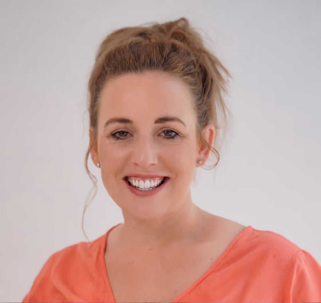 Smiling woman with brown hair in a ponytail wearing a coral top against a plain background.
