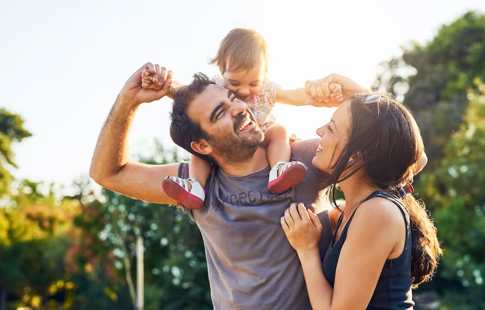 Smiling family outdoors: father carrying toddler on shoulders, mother holding father's arm, all joyful.