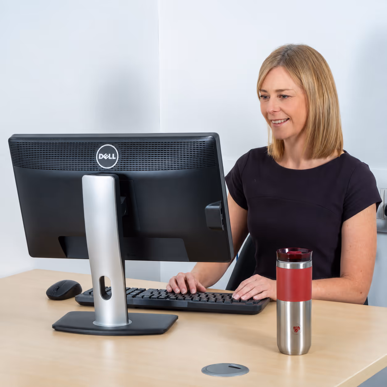 Smiling woman typing on keyboard at desk with Dell monitor and red stainless steel tumbler.