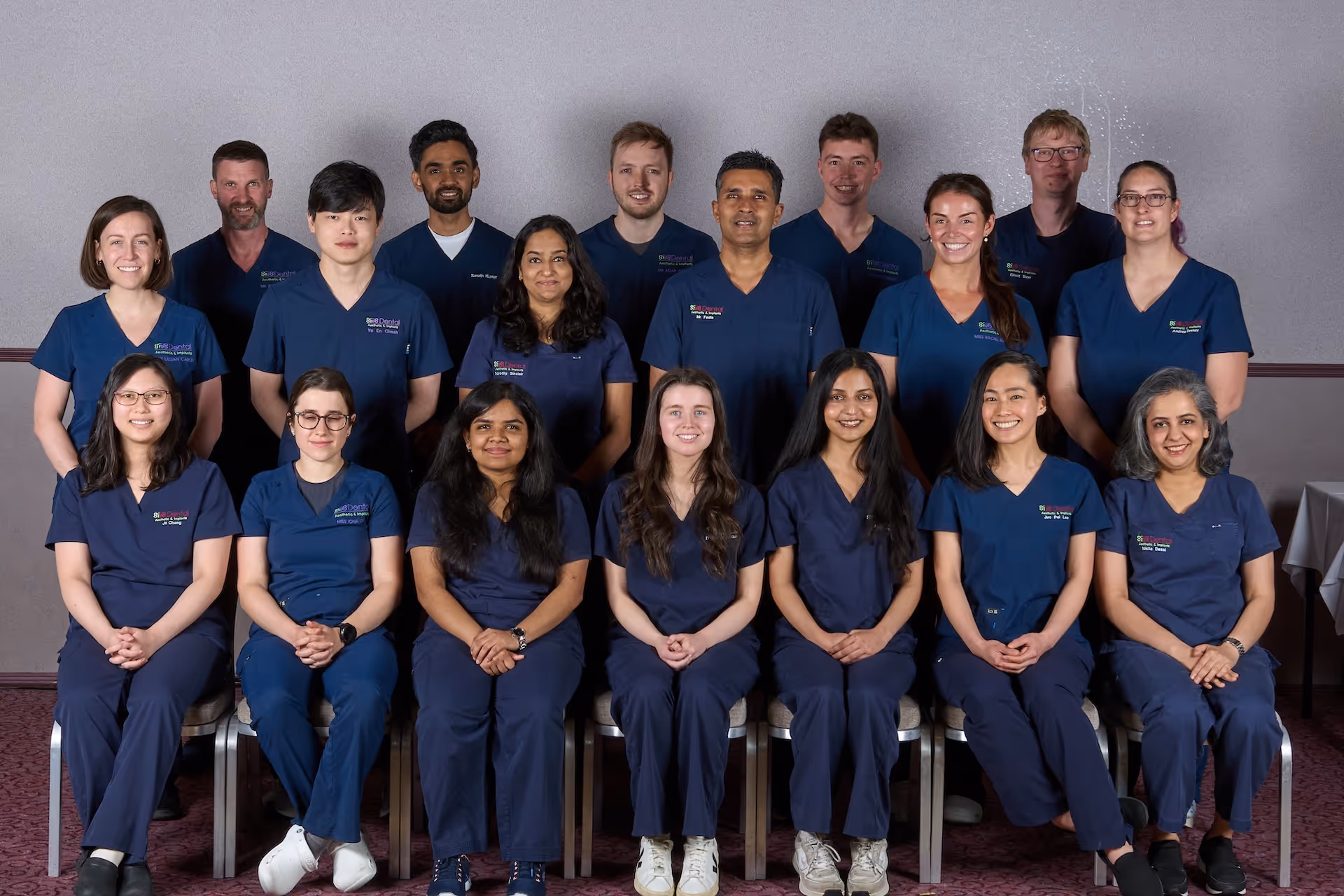 Group portrait of 18 diverse healthcare professionals in navy blue scrubs posing indoors.