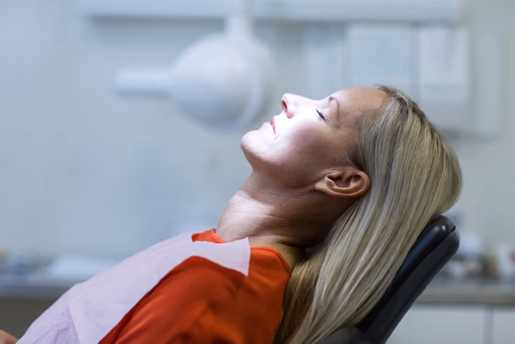 Relaxed woman reclined in a dental chair with eyes closed under bright dental light.