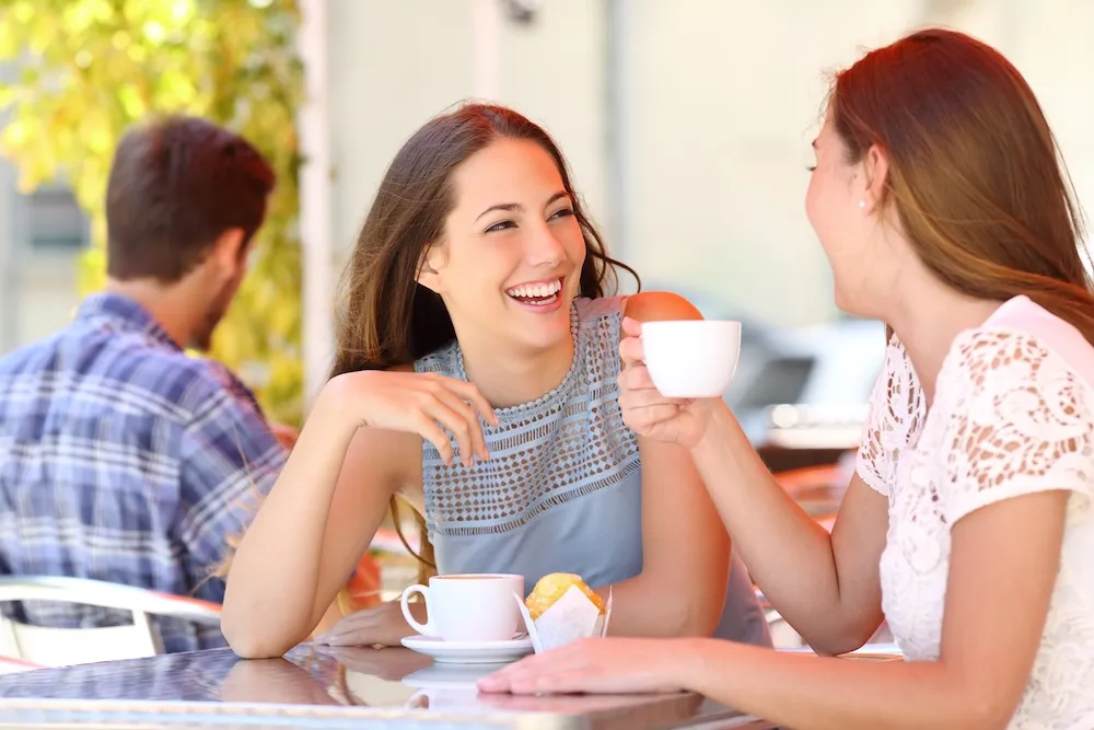 Two women smiling and sharing coffee at an outdoor cafe table with a muffin.